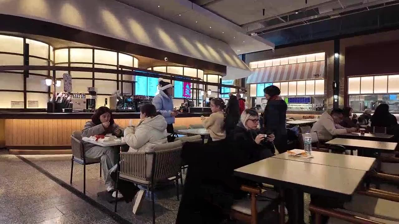 A busy restaurant at service: diners at every table in the foreground, people queueing at a long serving counter behind them.
