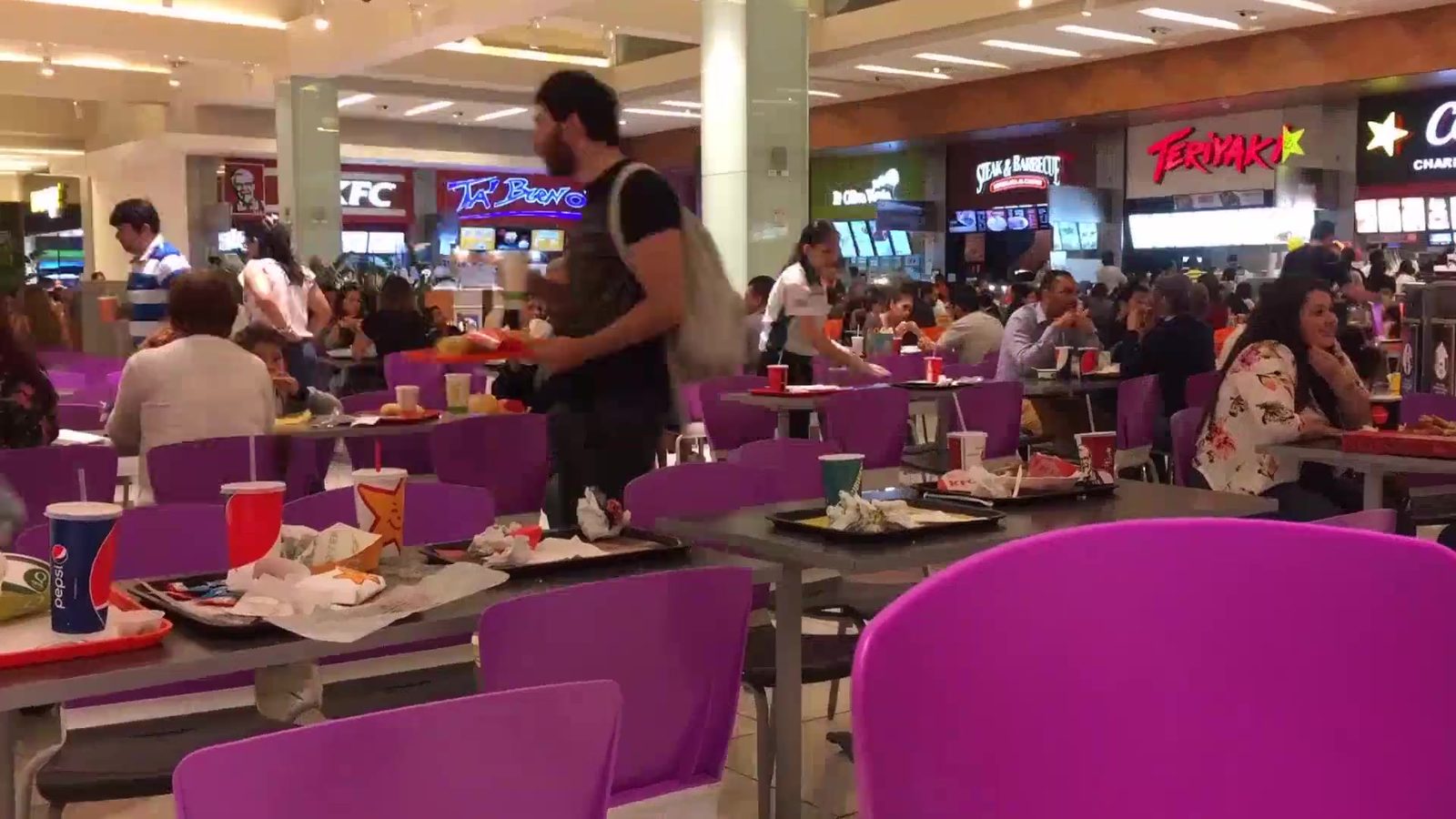 A busy food court at lunch: every table taken, diners eating at long tables with serving counters and lit menu screens along the back wall.